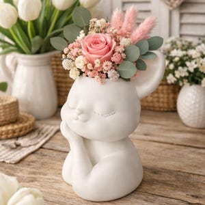 Könnte beinhalten: Eine weiße Keramikfigur einer schlafenden Maus, die als Blumenvase verwendet wird und mit einer rosa Rose, rosa flauschigem Gras und kleinen weißen und rosa Blüten gefüllt ist. Im Hintergrund befinden sich weitere weiße Vasen mit weißen Tulpen und Blumen.