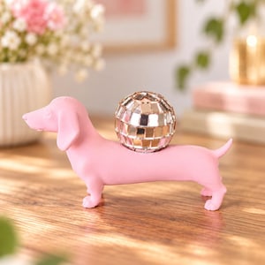 Könnte beinhalten: Eine matte, rosafarbene Dackel-Figur, die im Profil steht. Der Hund hat eine glatte, einfarbige Oberfläche und trägt auf dem Rücken zwischen den Schultern eine kleine, reflektierende silberne Discokugel mit quadratischen Spiegelkacheln.