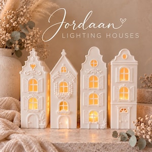 May include: Four white ceramic house-shaped lanterns with illuminated windows, featuring intricate architectural details like shutters and gables. The text Jordaan LIGHTING HOUSES is displayed above the structures in a white script and sans-serif font.