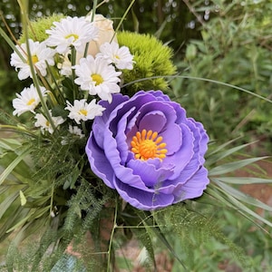 May include: A vibrant bouquet featuring a large, purple, layered flower with an orange center, surrounded by white daisies, green foliage, and other greenery. The arrangement is set against a blurred natural background.