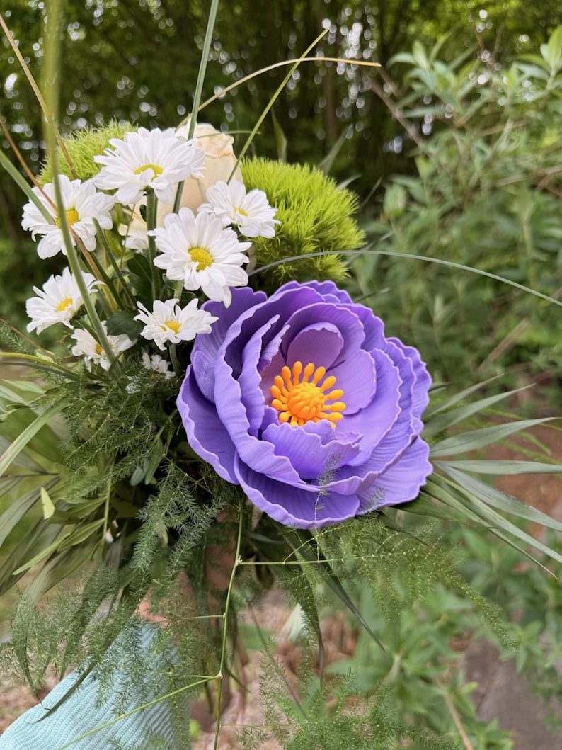 May include: A vibrant bouquet featuring a large, purple, layered flower with an orange center, surrounded by white daisies, green foliage, and other greenery. The arrangement is set against a blurred natural background.