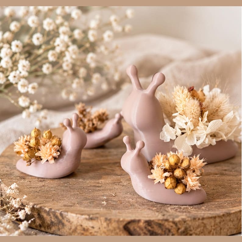 Op de afbeelding: Een verzameling van vier gladde, mauvekleurige slakvormige plantenbakken van verschillende groottes. Elk slakfiguur heeft een holle schelpruimte gevuld met een arrangement van gedroogde bloemen, waaronder kleine witte bloesems en zaaddozen.