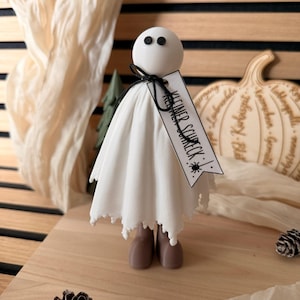 Könnte beinhalten: Eine weiße Geisterfigur mit rundem Kopf, zwei schwarzen Punktaugen und einem drapierten weißen Stoffkörper. Eine schwarze Schnur hält ein weißes Etikett um den Hals mit der Aufschrift "KLEINER SCHRECK" und einer kleinen schwarzen Spinne.