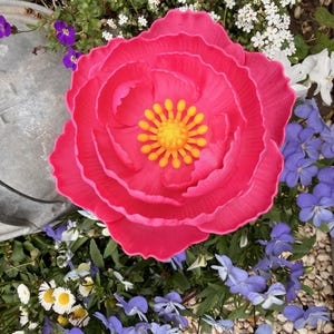 May include: A vibrant, large, pink artificial flower with a yellow center. The flower is surrounded by various small flowers in shades of white, purple, and blue, set against a backdrop of green foliage and small pebbles. The overall theme is a garden setting.