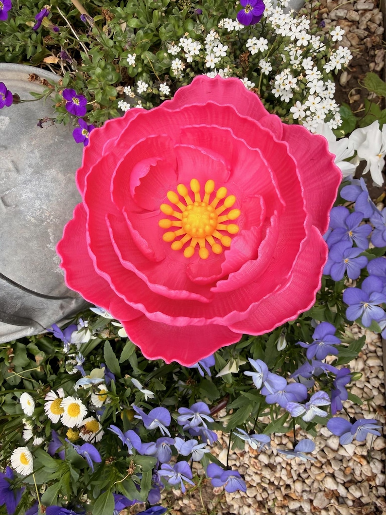 May include: A vibrant, large, pink artificial flower with a yellow center. The flower is surrounded by various small flowers in shades of white, purple, and blue, set against a backdrop of green foliage and small pebbles. The overall theme is a garden setting.