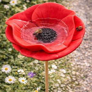 Könnte beinhalten: Ein leuchtend rotes, blütenförmiges Vogelbad mit dunkler Mitte, gefüllt mit Wasser. Eine Biene und ein Marienkäfer sitzen auf der Blüte. Das Vogelbad ist auf einem Holzpfahl montiert, vor einem Hintergrund aus Gänseblümchen und Grün.
