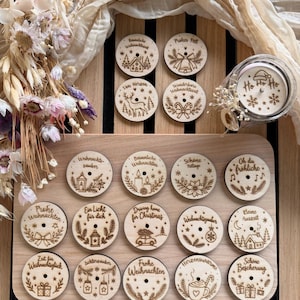 Könnte beinhalten: Eine Sammlung runder Holzscheiben mit lasergravierten weihnachtlichen Illustrationen und deutschen Texten wie Frohe Weihnachten, Besinnliche Weihnachten und Ho Ho Ho, die auf einem hellen Holzbrett mit einem mittigen Loch angeordnet sind.