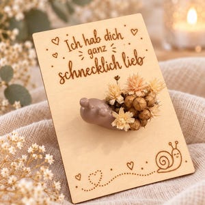 Op de afbeelding: Een rechthoekige houten kaart met de lasergegraveerde tekst Ich hab dich ganz schnecklich lieb, kleine hartjes en een slakillustratie. Een gladde, lichtbruine slakfiguur met een huisje van gedroogde beige bloemen is bevestigd.