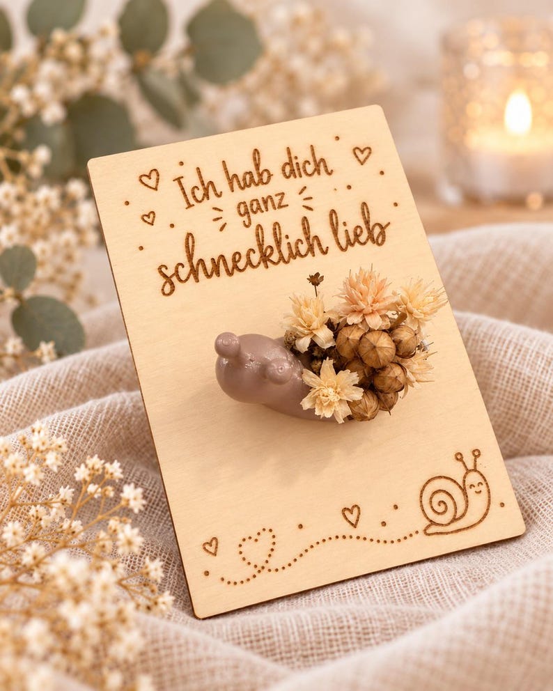 Op de afbeelding: Een rechthoekige houten kaart met de lasergegraveerde tekst Ich hab dich ganz schnecklich lieb, kleine hartjes en een slakillustratie. Een gladde, lichtbruine slakfiguur met een huisje van gedroogde beige bloemen is bevestigd.