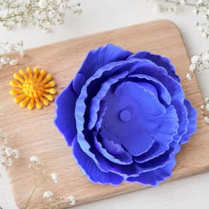 May include: A vibrant blue peony flower and a small yellow flower are displayed on a wooden cutting board. Delicate white flowers surround the board. The peony has detailed petals, and the yellow flower has a sunburst design.
