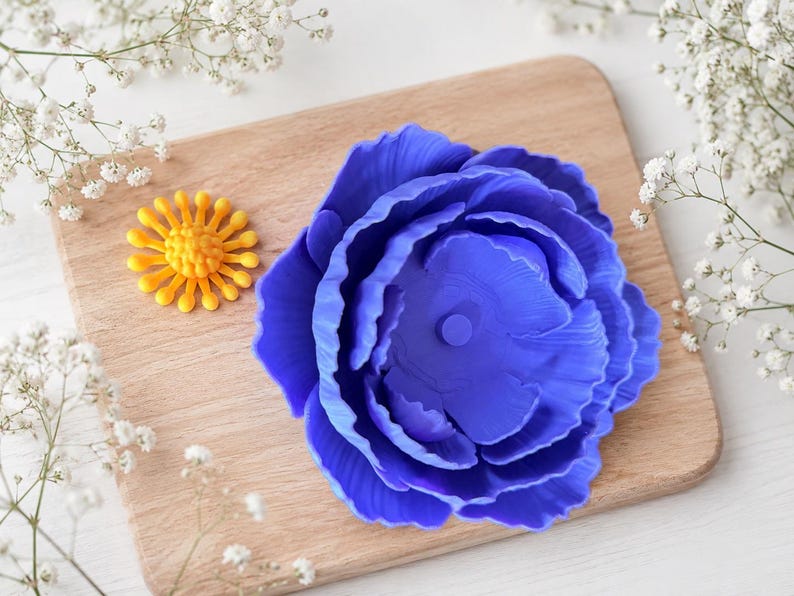May include: A vibrant blue peony flower and a small yellow flower are displayed on a wooden cutting board. Delicate white flowers surround the board. The peony has detailed petals, and the yellow flower has a sunburst design.