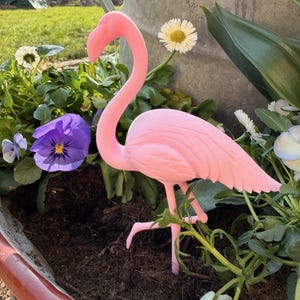 May include: A pink flamingo garden ornament stands amidst colorful flowers and greenery in a decorative planter. The flamingo has a long, curved neck and is posed with one leg raised, adding a touch of whimsy to the outdoor setting.