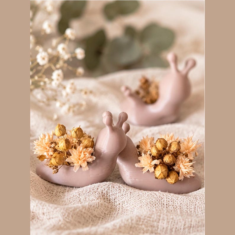 Op de afbeelding: Drie gladde, matte, mauvekleurige plantenbakjes in de vorm van een slak. Elk slakfiguur heeft een holle opening op de rug, gevuld met een bosje gedroogde, lichtbeige en lichtbruine bloemen. De slaksculpturen zijn in een groep geplaatst.