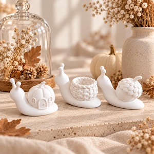 May include: Three white snail figurines with distinct shells. The first snail has a pumpkin-shaped shell with a door and windows. The second snail features a woven basket shell. The third snail carries a textured acorn-shaped shell on its back.