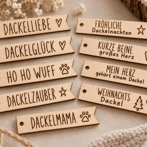 Könnte beinhalten: Neun rechteckige Anhänger aus hellem Holz mit einem Loch auf der linken Seite und einem spitz zulaufenden Ende rechts. Jeder Anhänger ist mit gelaserten deutschen Texten und Symbolen wie Herzen, Sternen, Pfotenabdrücken und einem Tannenbaum versehen.