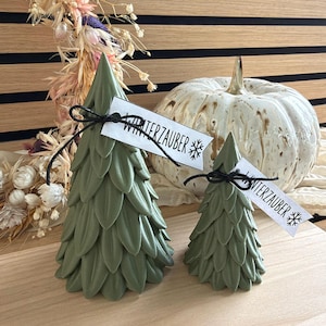 Pode incluir: Duas velas em forma de árvore na cor verde sálvia com texturas de folhas em camadas. Cada vela está amarrada com um fio preto fino que segura uma etiqueta de papel branca com o texto "WINTERZAUBER" e um ícone de floco de neve impresso.
