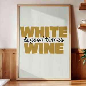 Peut inclure: Une affiche encadrée avec les mots "WHITE & good times WINE" en lettres dorées et noires. L'affiche est exposée sur un comptoir en bois dans une cuisine. Un évier blanc et des étagères avec de la vaisselle sont visibles.
