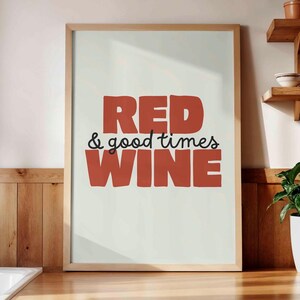 Peut inclure: Une affiche encadrée avec les mots "RED & good times WINE" en lettres rouges et noires. L'affiche est exposée sur un comptoir en bois dans une cuisine, avec des étagères et une plante en arrière-plan.