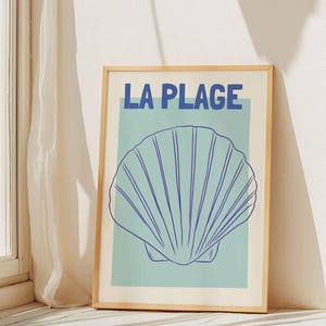 Peut inclure: Une affiche encadrée avec le texte "LA PLAGE" en bleu au-dessus d'une illustration de coquillage bleu sur fond bleu clair. L'affiche est dans un cadre en bois et est posée contre un mur blanc près d'une fenêtre avec vue sur la mer.