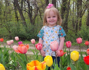 Puede incluir: Una niña pequeña con una camisa estampada en azul y blanco con un lazo rosa en el pelo está de pie en un campo de tulipanes coloridos. Los tulipanes son de varios tonos de rosa, amarillo, naranja y rojo.