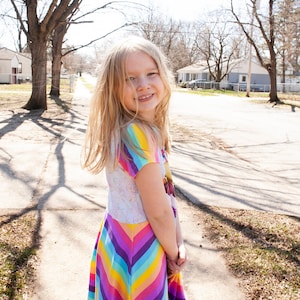 May include: A young girl with long blonde hair smiles at the camera while wearing a rainbow striped dress. She is standing on a sidewalk in front of a row of houses.