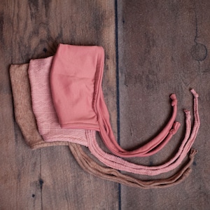 Pode incluir: Três gorros de recém-nascido em tons de rosa e bege são dispostos sobre uma superfície de madeira. Os gorros têm uma variedade de texturas, incluindo tecidos de malha e lisos. Cada gorro tem laços longos.