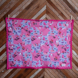 以下が含まれることがあります： ユニコーンと蝶の模様が施されたピンクとパープルのパッチワークキルト。キルトはピンクの縁取りで飾られています。