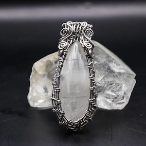 Puede incluir: Un colgante de cristal de cuarzo transparente con un alambre de plata. El colgante tiene forma de lágrima y se encuentra sobre una piedra rugosa de color claro. El alambre tiene detalles intrincados en la parte superior.