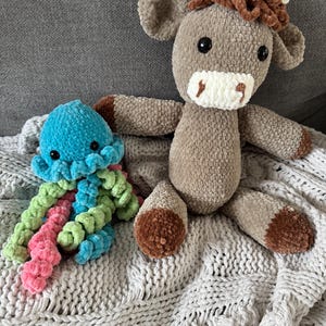 May include: Two handmade crochet plush toys: a light brown cow with white horns and a blue octopus with pink and green tentacles. The cow has a white muzzle and brown hooves. Both are on a gray knitted blanket.