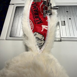 May include: A close-up of a sewing machine stitching a white plush fabric with a red and white patterned fabric. The red fabric has the text "ALABAMA" and a bulldog graphic. The sewing machine has measurements in inches.