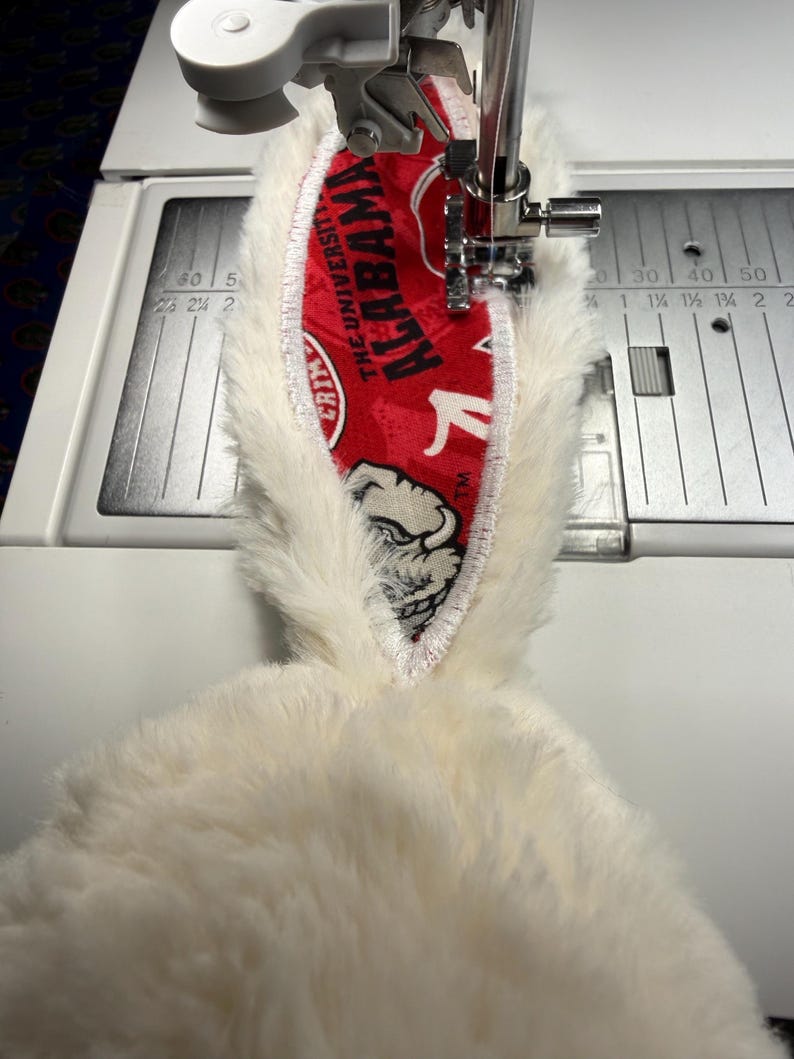 May include: A close-up of a sewing machine stitching a white plush fabric with a red and white patterned fabric. The red fabric has the text "ALABAMA" and a bulldog graphic. The sewing machine has measurements in inches.