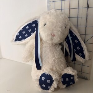 May include: A plush, cream-colored bunny with navy blue patterned accents on the ears and feet. The bunny has a pink nose and is adorned with navy and light blue ribbons. The bunny measures approximately 10 inches in height.