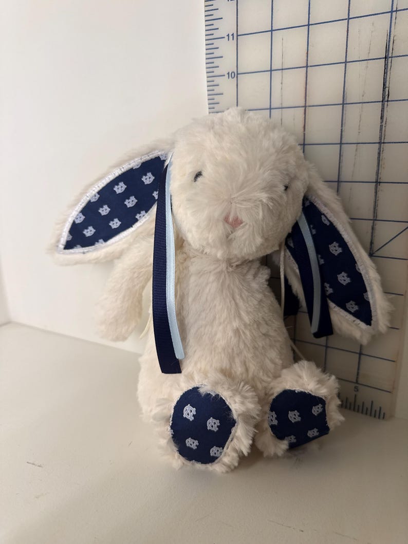 May include: A plush, cream-colored bunny with navy blue patterned accents on the ears and feet. The bunny has a pink nose and is adorned with navy and light blue ribbons. The bunny measures approximately 10 inches in height.