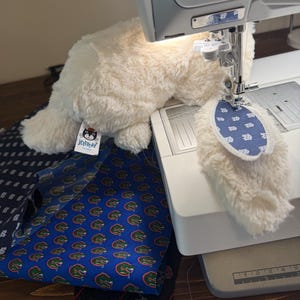 May include: A cream-colored plush cat toy with a blue and white patterned tail is being sewn on a sewing machine. Several patterned fabrics, including a blue fabric with green alligators, are visible.