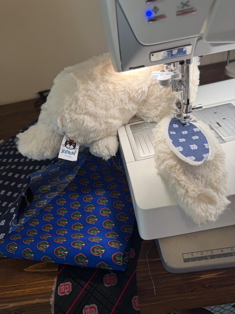 May include: A cream-colored plush cat toy with a blue and white patterned tail is being sewn on a sewing machine. Several patterned fabrics, including a blue fabric with green alligators, are visible.