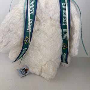 May include: A fluffy, off-white stuffed animal with two ribbons attached to its back. The ribbons are white, teal, and dark green, with the text "UNCW SEAHAWKS" printed on them. A small tag with a cat logo is visible.