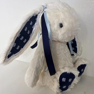May include: A plush white bunny rabbit with navy blue accents. The bunny's ears and feet feature a navy blue fabric with a repeating pattern. Navy and light blue ribbons are tied to the ears. A soft, cuddly toy.