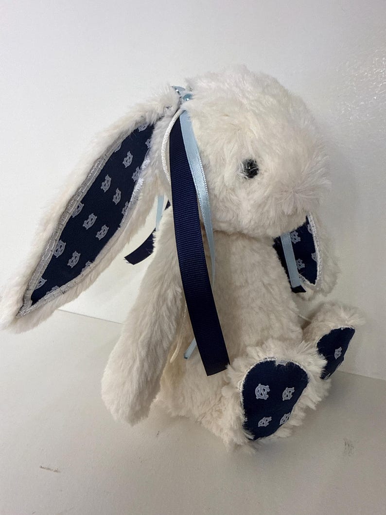 May include: A plush white bunny rabbit with navy blue accents. The bunny's ears and feet feature a navy blue fabric with a repeating pattern. Navy and light blue ribbons are tied to the ears. A soft, cuddly toy.