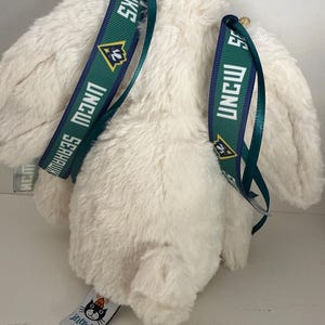 May include: A cream-colored plush bunny with green and navy blue straps featuring the text "UNCW Seahawks". The bunny has long ears and a small tag with the "Jellycat London" logo.