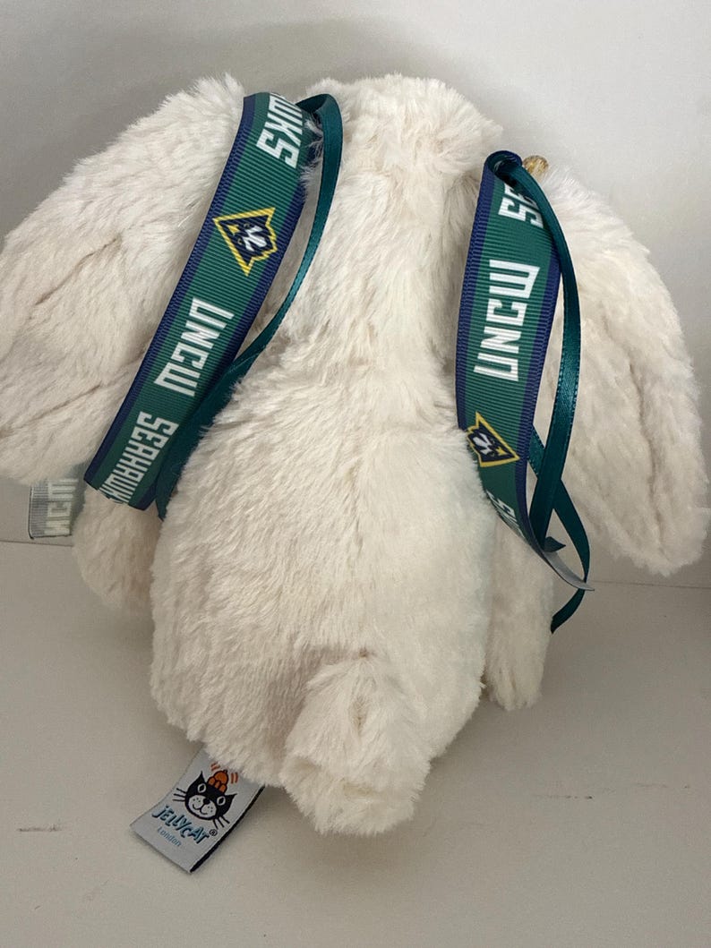May include: A cream-colored plush bunny with green and navy blue straps featuring the text "UNCW Seahawks". The bunny has long ears and a small tag with the "Jellycat London" logo.