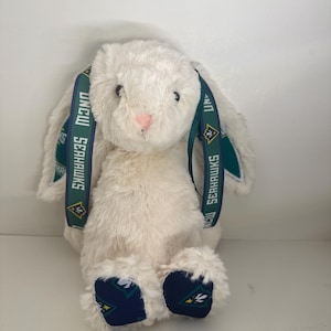 May include: A plush white bunny with long ears and a pink nose. The bunny has a green and navy blue ribbon around its ears with the text "UNCW SEAHAWKS". The bunny's feet are navy blue with a green and white design.