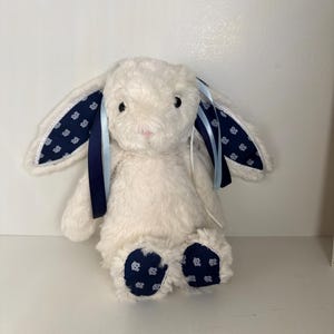 May include: A plush white bunny rabbit with long ears and a pink nose. The inner ears and foot pads feature a navy blue fabric with a white floral pattern. Navy and light blue ribbons are tied to the ears.