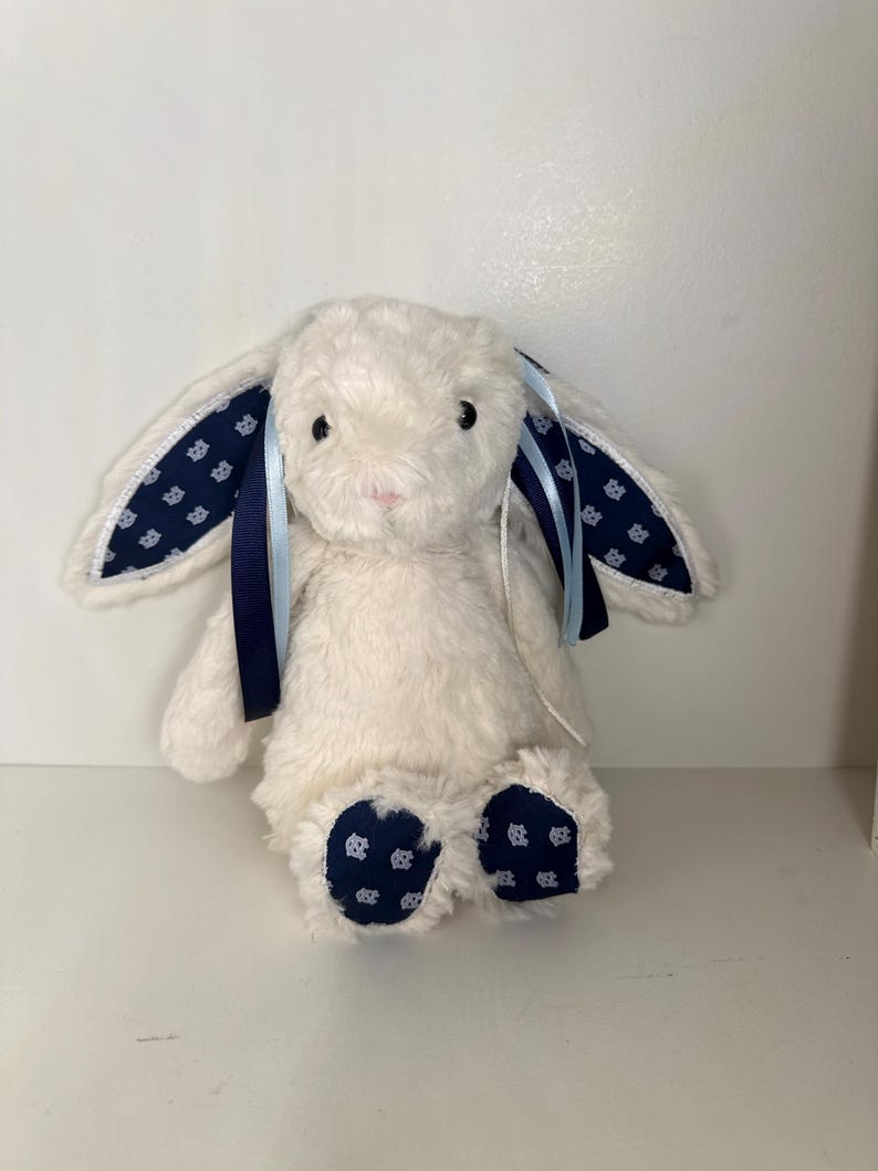May include: A plush white bunny rabbit with long ears and a pink nose. The inner ears and foot pads feature a navy blue fabric with a white floral pattern. Navy and light blue ribbons are tied to the ears.