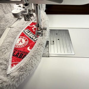 May include: A sewing machine stitching a red and white patch with the text "ALABAMA CRIMSON TIDE" onto a gray, fluffy fabric. The patch features the letter "A" and the words "THE UNIVERSITY OF ALABAMA". The machine's needle is in action.
