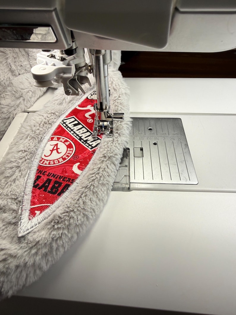 May include: A sewing machine stitching a red and white patch with the text "ALABAMA CRIMSON TIDE" onto a gray, fluffy fabric. The patch features the letter "A" and the words "THE UNIVERSITY OF ALABAMA". The machine's needle is in action.