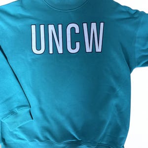 May include: Teal sweatshirt with the white letters "UNCW" across the chest. The letters have a black outline. The sweatshirt has a mock neck and ribbed cuffs and hem. The garment is hanging on a pink hanger.