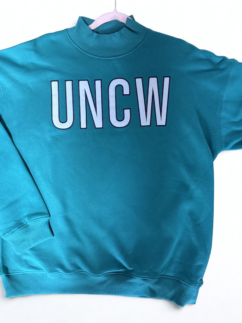 May include: Teal sweatshirt with the white letters "UNCW" across the chest. The letters have a black outline. The sweatshirt has a mock neck and ribbed cuffs and hem. The garment is hanging on a pink hanger.