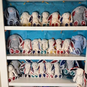 May include: A collection of plush bunny rabbits in various colors, arranged on shelves. The bunnies have long floppy ears and are decorated with ribbons and patches. Some bunnies are gray, others are white, and some have patterned ears.