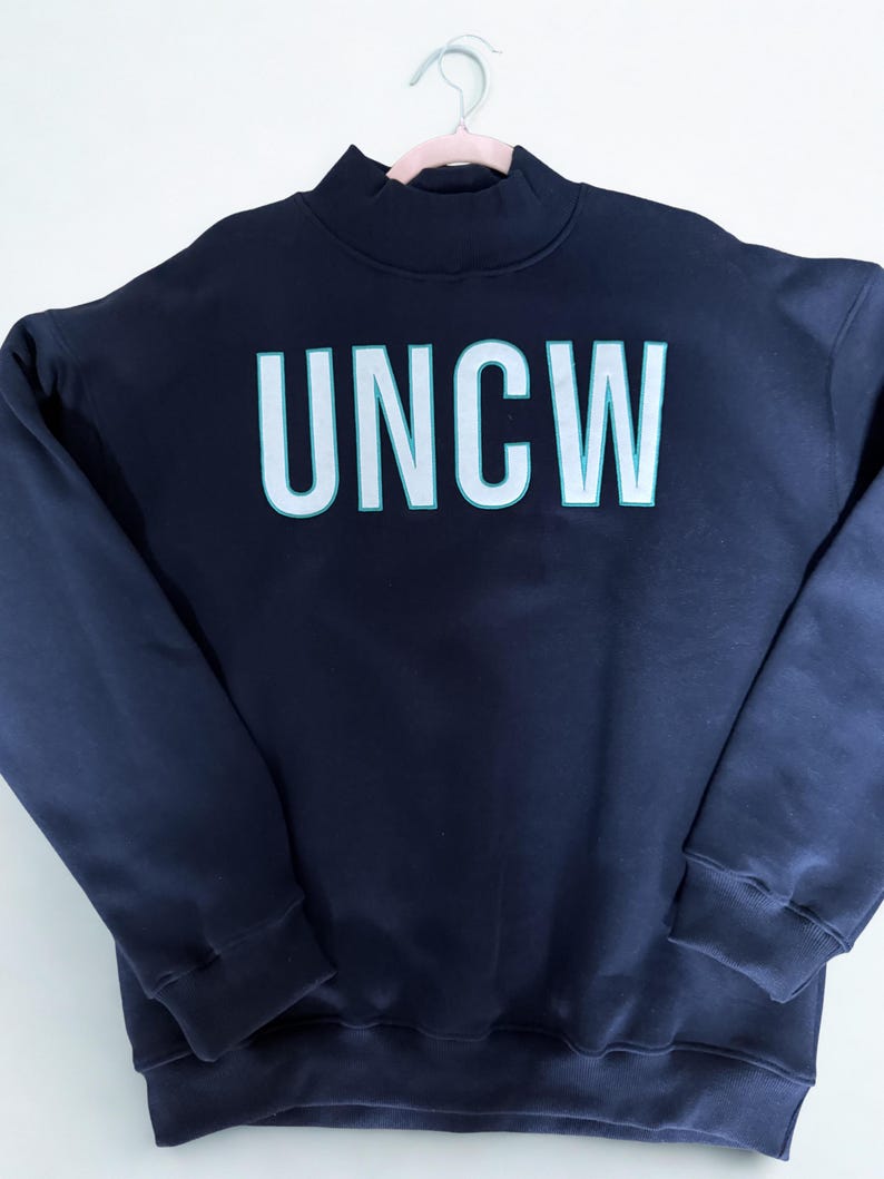 May include: A navy blue sweatshirt with the letters "UNCW" in large, white, block letters with a teal outline. The sweatshirt has a mock neck and long sleeves with ribbed cuffs. The garment is hanging on a pink hanger.