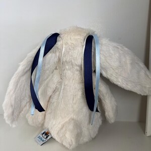May include: A plush, cream-colored bunny toy with long ears and a fluffy texture. The bunny has dark blue and light blue ribbons tied to its ears. A small tag with a logo is attached to the bunny's body.